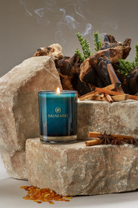 Single 10.5oz Balneario Luxury Soy-Coconut Candle in teal glassware, with a flame on stones, surrounded by Hammam-scented ingredients.