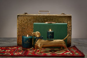 Two Balneario candles atop a bronze dog sculpture, against a gold suitcase backdrop on a Moroccan rug.