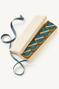 Balneario fragrance discovery set displayed in a gift box and ribbon with 5 gold tins. Each tin has a teal label with gold text describing each fragrance.