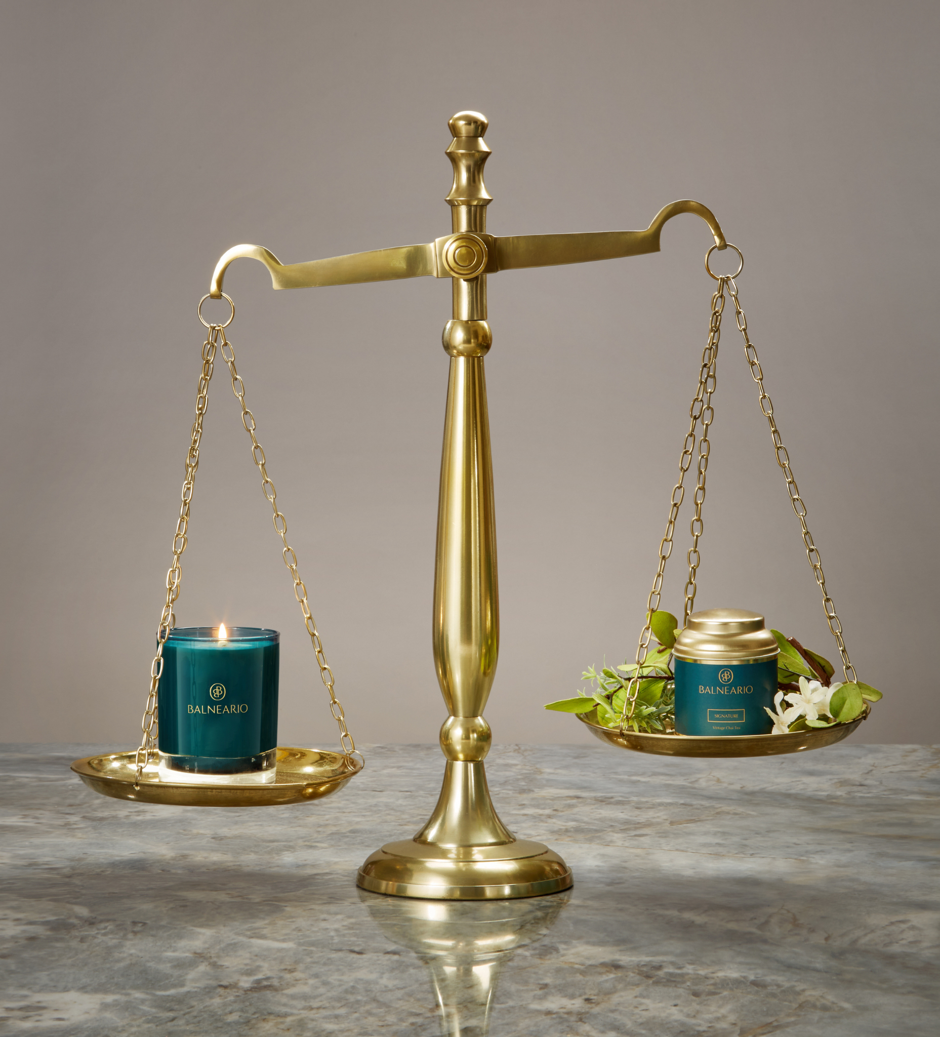 Balneario 10.5 ounce candle in a teal vessel balance on a gold scale along with a Balneario tea in a gold tin with leaves. 