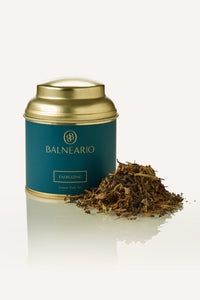 Balneario Energizing Wellness Tea in a teal tin with a gold lid, alongside a fragrant pile of energizing loose tea leaves. 