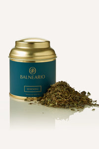 Balneario Refreshing Wellness Tea in a teal tin with a gold lid, alongside a fragrant pile of refreshing loose tea leaves.