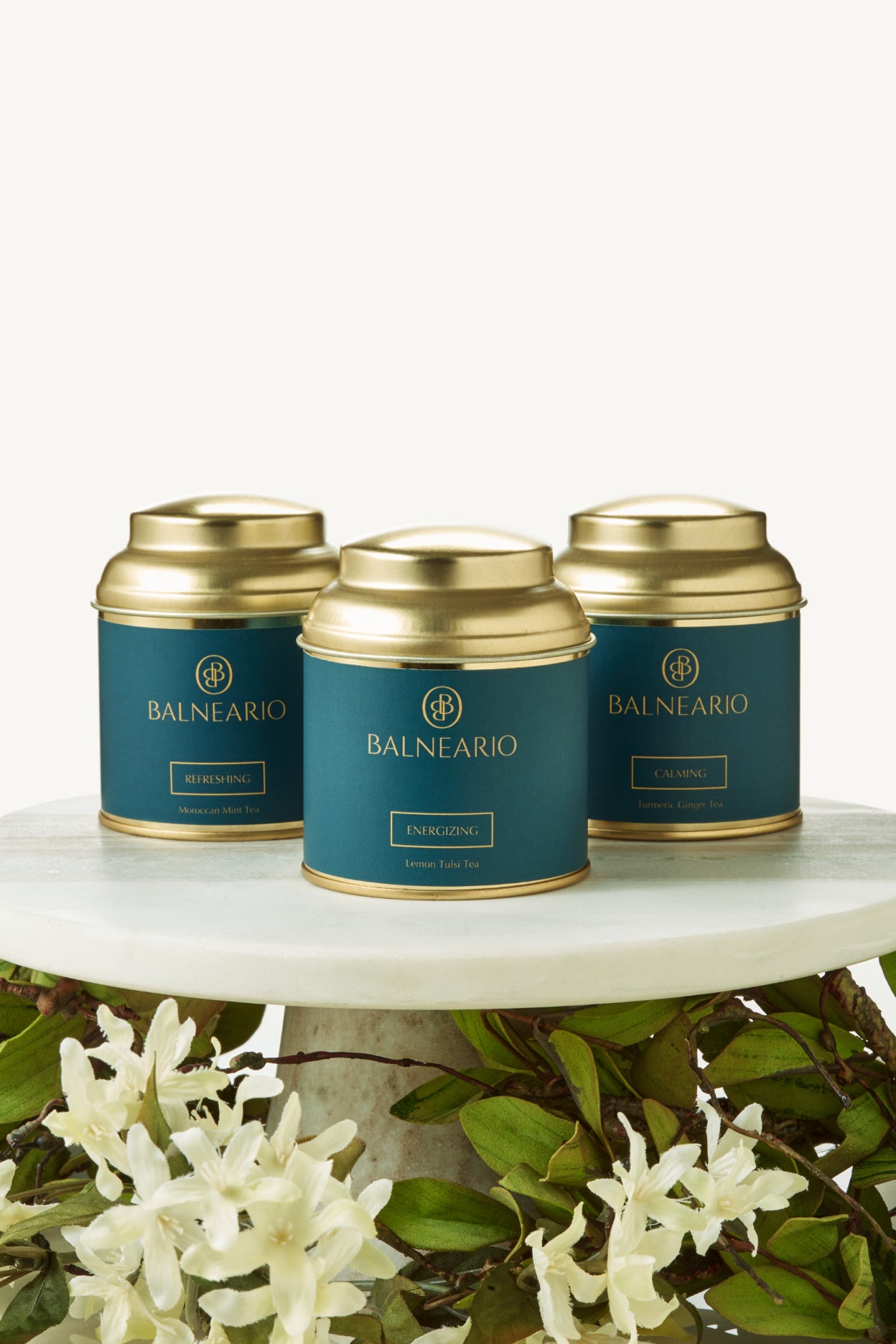 Balneario Wellness Tea Trio in teal tins with gold lids on a cake stand pedestal surrounded by flowers.  